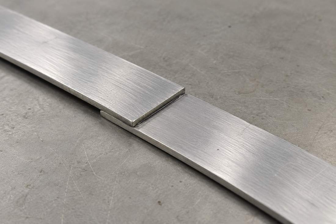 Zwei flache Metallstreifen sind an ihren Enden mit einer dünnen Klebfuge überlappt.